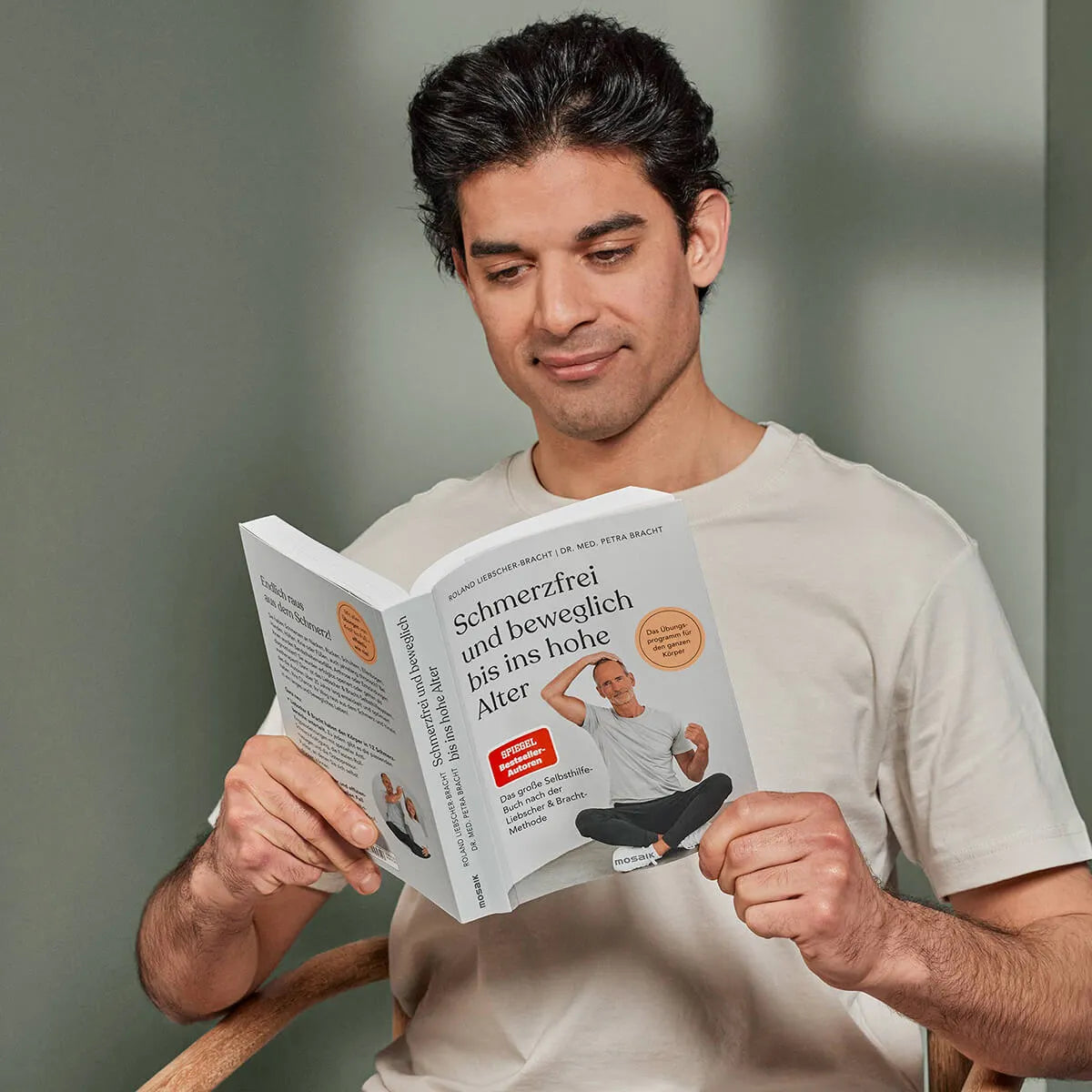 Ein dunkelhaariger Mann in einem beigen T-Shirt sitzt auf einem Stuhl und liest lächelnd "Schmerzfrei und beweglich bis ins hohe Alter" von Liebscher & Bracht in einem sanft beleuchteten Raum mit grünen Wänden.