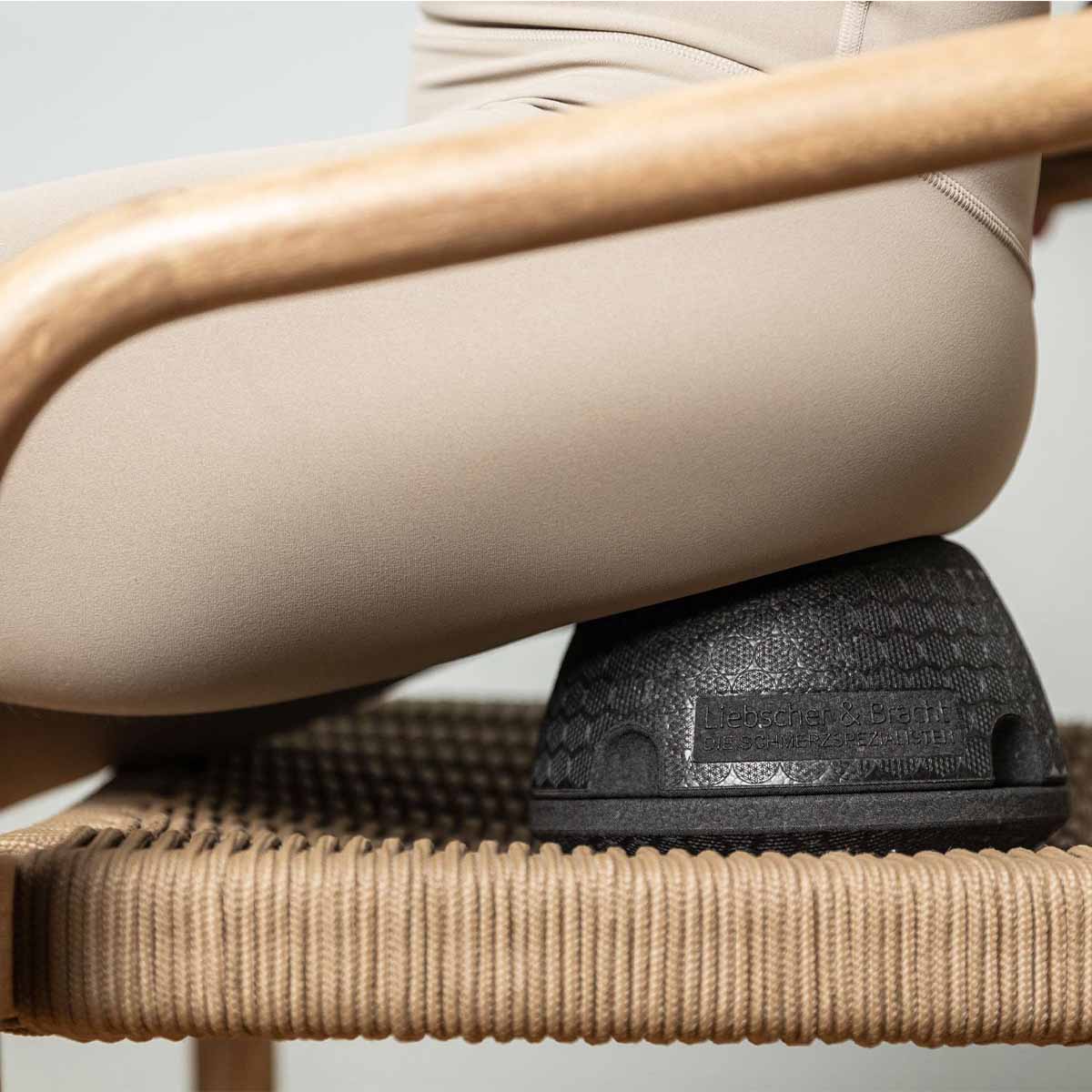 Eine Person in beigen Leggings sitzt auf dem ISG-Ischias-Retter von Liebscher & Bracht, einem schwarzen kuppelförmigen Kissen auf einem geflochtenen Stuhl, um den Hüftbereich zu unterstützen und die Beweglichkeit zu verbessern.