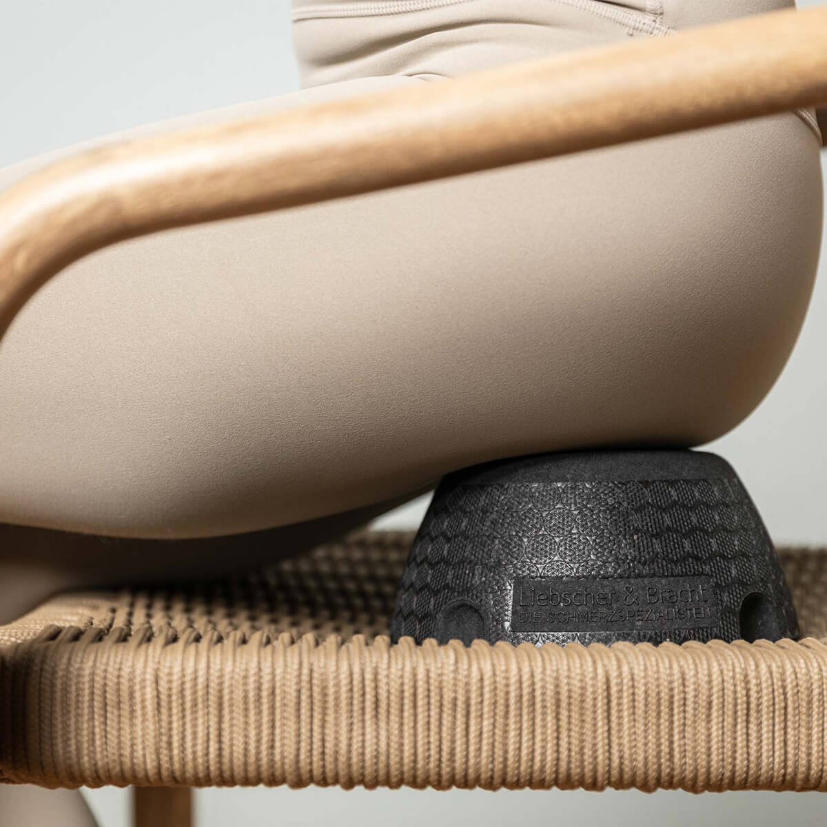 Eine Person in beigen Leggings sitzt auf einem geflochtenen Stuhl, zwischen ihr und der Sitzfläche des Stuhls befindet sich ein schwarzes, strukturiertes Kissen oder ein Stützobjekt.