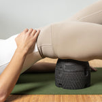 Eine Person benutzt den schwarzen Der ISG-Ischias-Retter von Liebscher & Bracht auf einer grünen Matte im Hüft- oder Gesäßbereich, trägt beige Leggings und ein weißes Oberteil und übt eine Übung zur Verbesserung der Beweglichkeit.