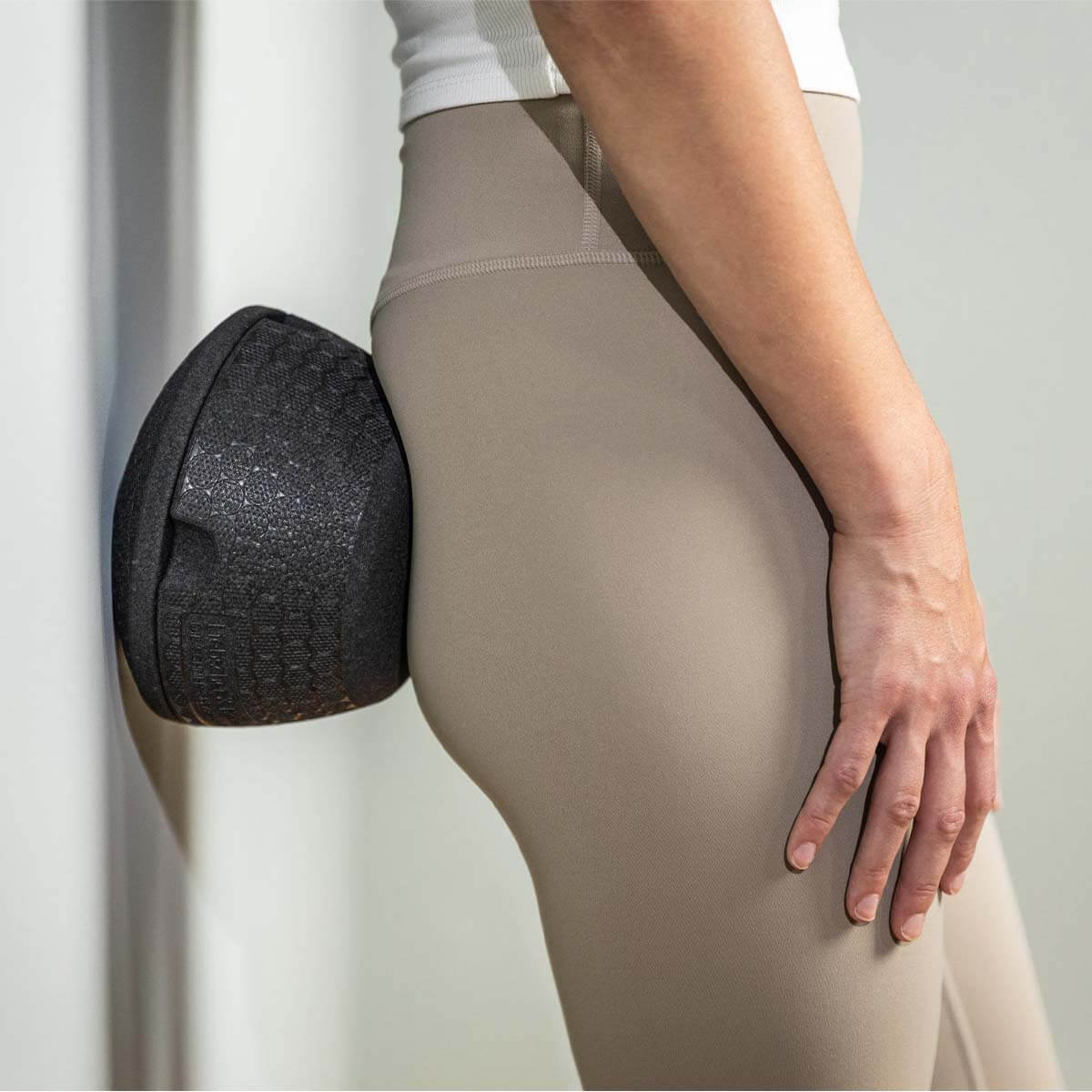 Eine Person benutzt die schwarze Liebscher & Bracht ISG-Ischias-Retter Schaumstoffrolle gegen eine Wand, um ihren Hüft- und Gesäßbereich zu massieren und die Beweglichkeit zu verbessern.