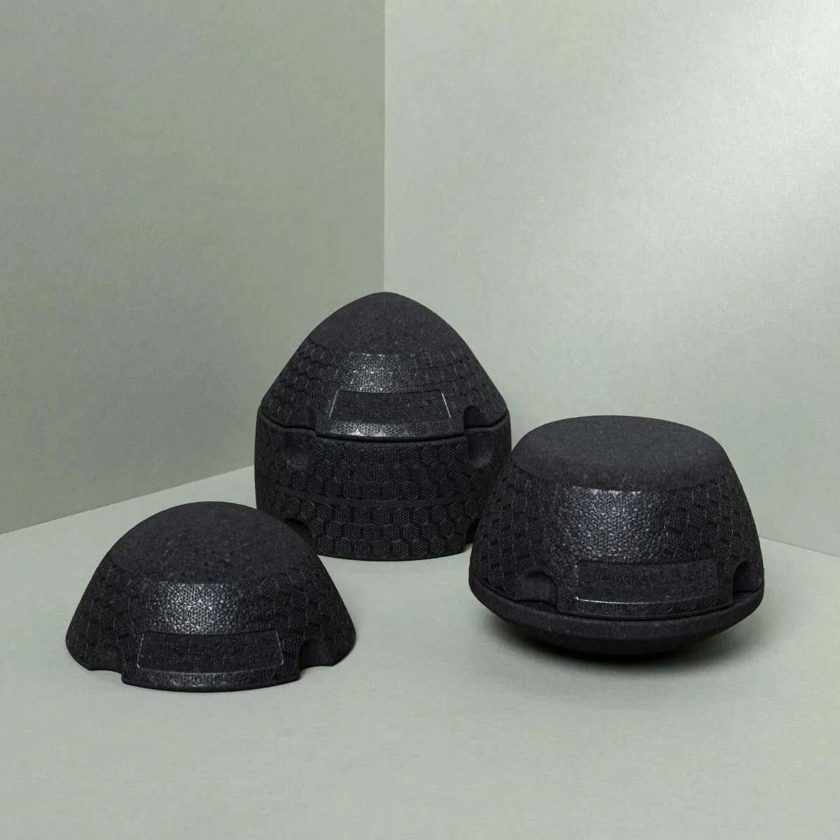 Drei schwarze Der ISG-Ischias-Retter von Liebscher & Bracht, ideal zur Verbesserung der Beweglichkeit im Hüft- oder Gesäßbereich, stehen in Dreiecksformation auf einer hellgrauen Fläche vor einem passenden Hintergrund.