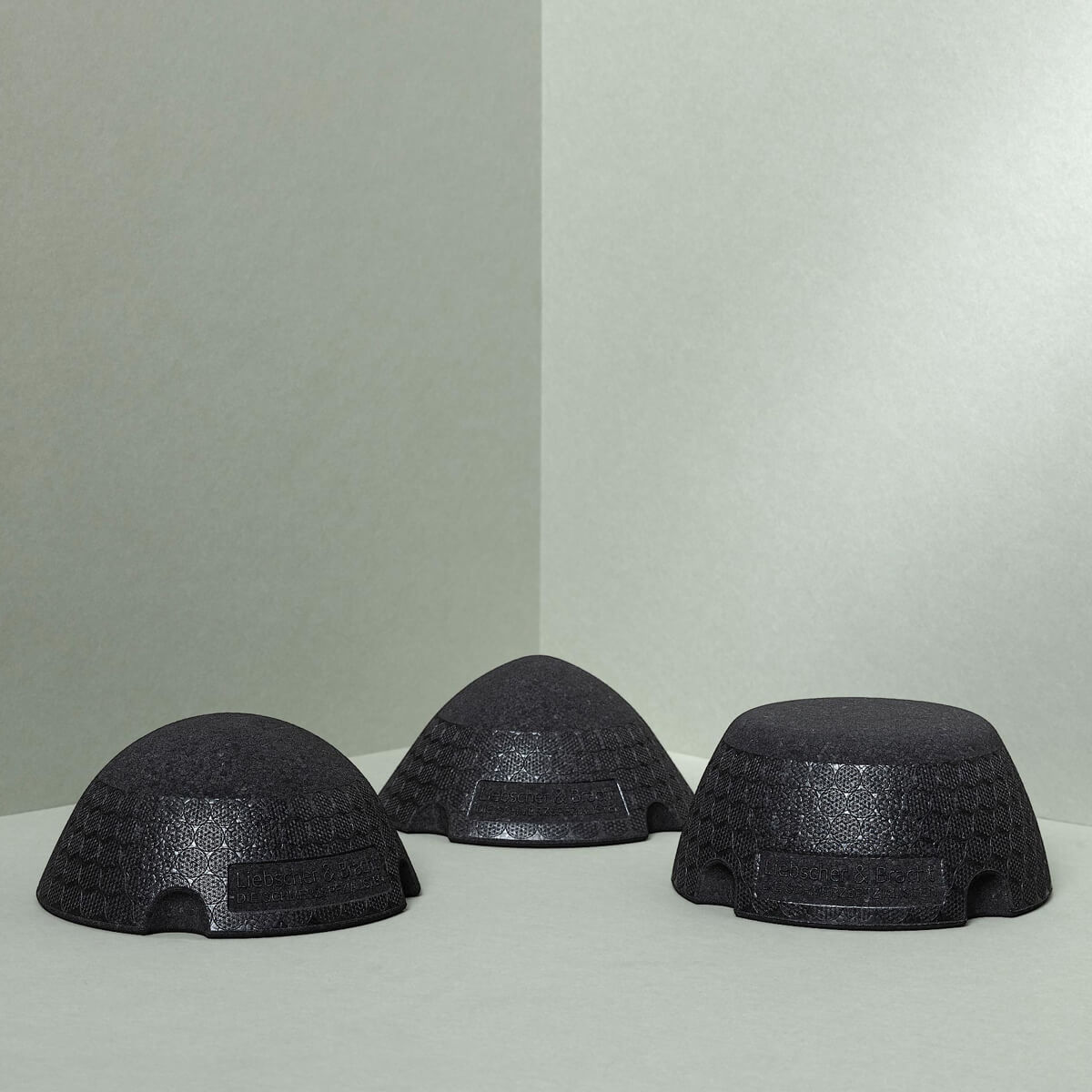 Drei schwarz strukturierte, kuppelförmige Liebscher & Bracht Der ISG-Ischias-Retter-Objekte ruhen auf einer hellgrauen Fläche in einer hellgrauen Ecke, jeweils in unterschiedlichen Winkeln angeordnet. Eingerückte rechteckige Abschnitte deuten ihre Beweglichkeits-Trainingsfunktion an.