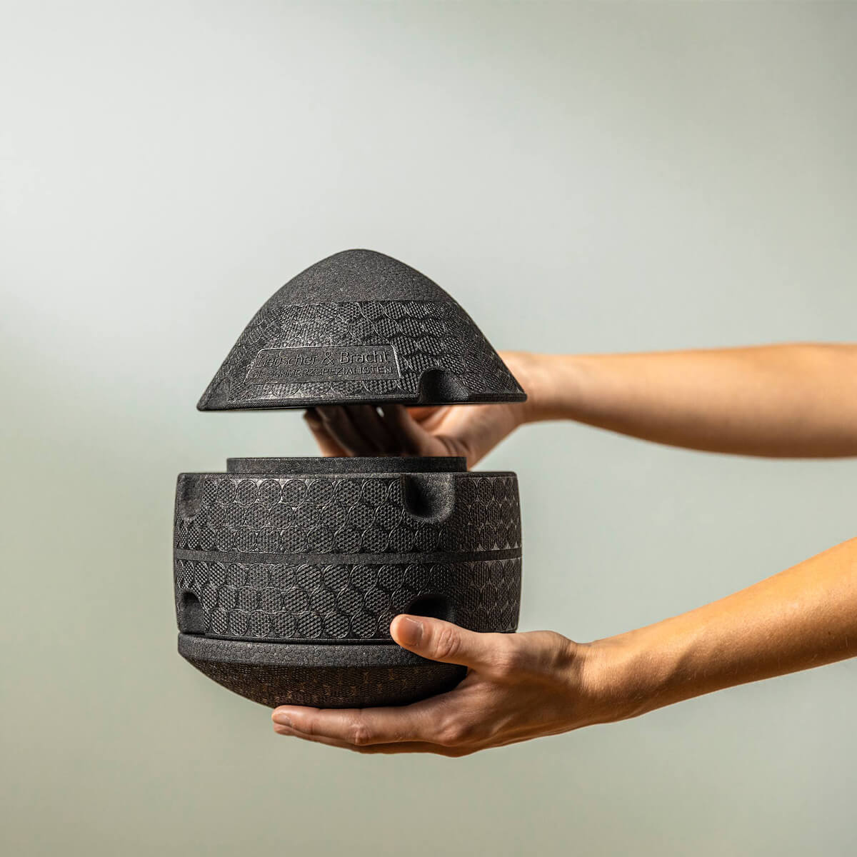 Zwei Hände halten den strukturierten, schwarzen ISG-Ischias-Retter von Liebscher & Bracht. Der geometrisch gemusterte Deckel wird angehoben und gibt einen hohlen Innenraum frei - ideal zur Aufbewahrung von kleinem Fitnesszubehör für den Hüft- oder Gesäßbereich.