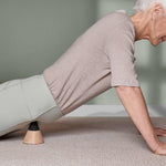 Ein älterer Erwachsener in einem beigen Oberteil und einer hellgrauen Hose führt Druckübungen im Schneidersitz mit dem Liebscher & Bracht Schmerzfrei-Drücker-Set unter den Hüften auf einem hellen Teppichboden vor einer grünen Wand durch.