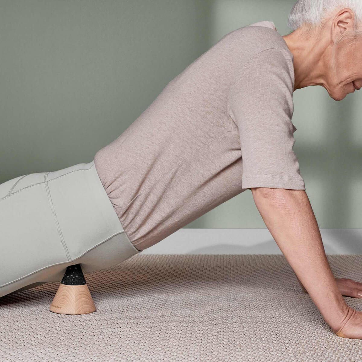 Ein älterer Erwachsener in einem beigen Oberteil und einer hellgrauen Hose führt Druckübungen im Schneidersitz mit dem Liebscher & Bracht Schmerzfrei-Drücker-Set unter den Hüften auf einem hellen Teppichboden vor einer grünen Wand durch.