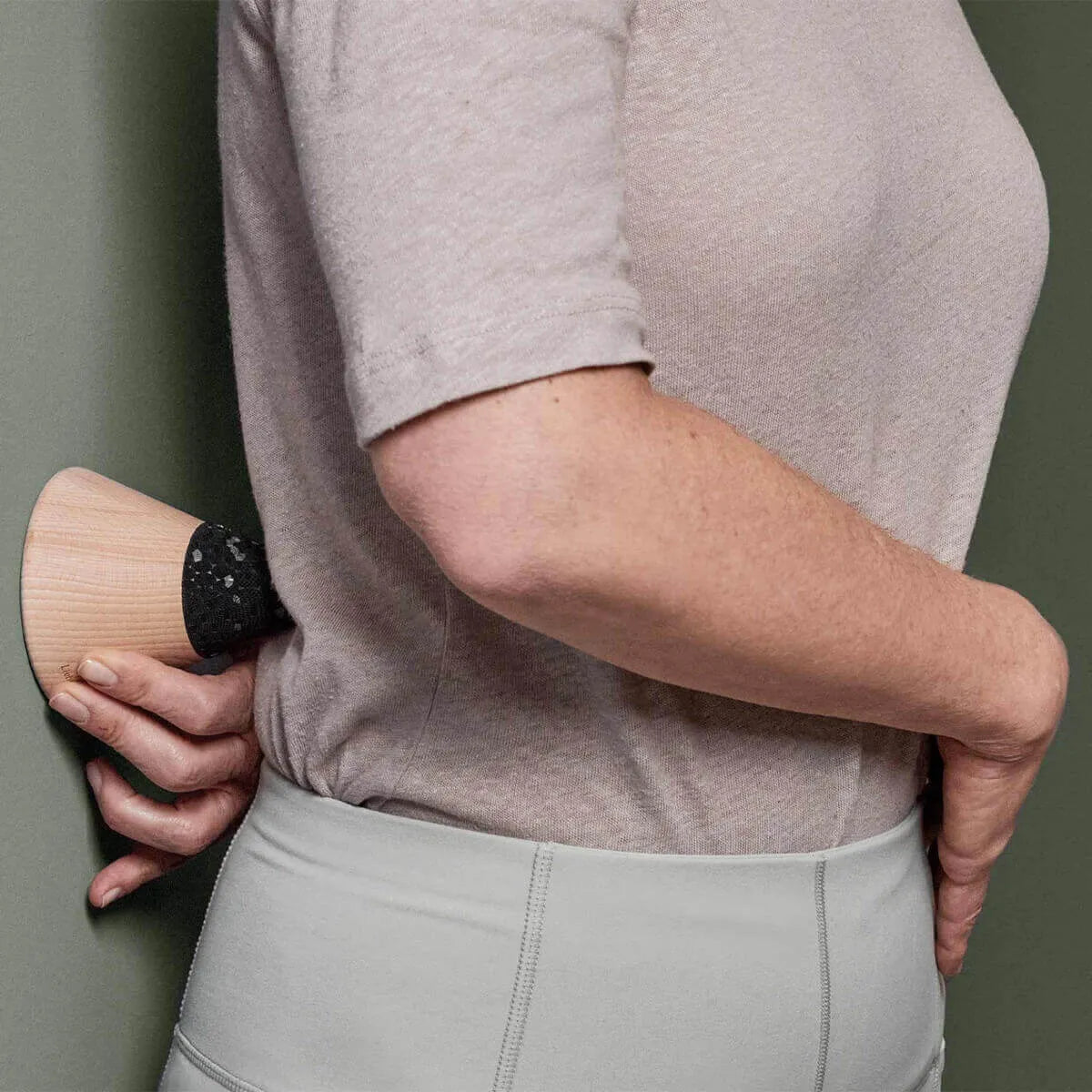 Eine Person in beigem Hemd und hellgrauer Hose setzt ein gebogenes Holzmassagegerät mit schwarzem Griff am unteren Rücken ein - ideal für das Programm "Rücken- & Bandscheibenschmerzen selbst behandeln" von Liebscher & Bracht.