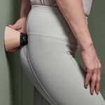 Eine Person in hellgrauen Leggings benutzt ein hölzernes Massagegerät mit schwarzem Griff aus dem Liebscher & Bracht Angebots-Paket und drückt es gegen ihre Hüfte, während sie an einer grünen Wand steht.