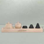 Das Aufbewahrungsbrett von Liebscher & Bracht fasst zwei unterschiedlich geformte Massagewerkzeuge aus hellem Holz und zwei schwarze, kegelförmige Massagesteine in einer Reihe, auf deren Holzsockel der Markenname aufgedruckt ist.