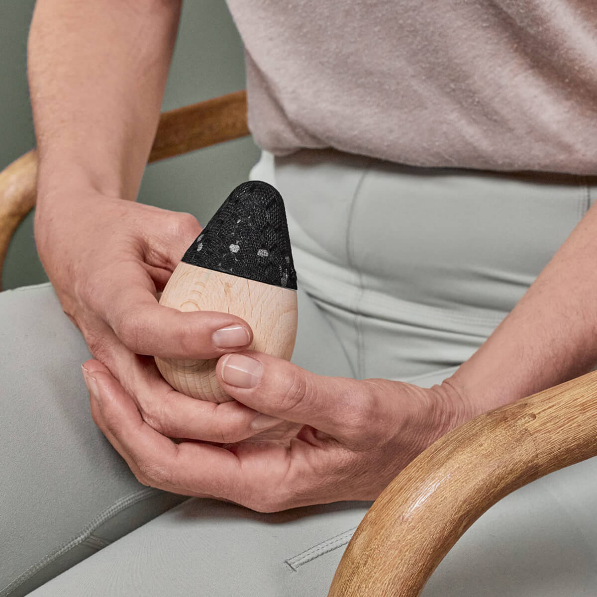 Auf einem Holzstuhl sitzend, hält eine Person das Liebscher & Bracht Schmerzfrei-Drücker-Set - ein eiförmiges Werkzeug mit schwarzer, strukturierter Oberfläche - ideal für Licht-Osteopressur und Druckübungen.