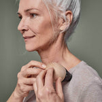 Eine ältere Frau mit grauem Haar trägt das Liebscher & Bracht Schmerzfrei-Drücker-Set auf der Schulter und lächelt sanft in einem hellbeigen Oberteil vor einem neutralen Hintergrund.