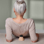 Eine grauhaarige Frau sitzt mit dem Gesicht zur Seite und stützt sich auf ein hölzernes und schwarzes Gerät aus dem Liebscher & Bracht-Angebots-Paket, das sie unter ihrem unteren Rücken auf einer strukturierten Oberfläche trägt und wahrscheinlich zur Unterstützung oder Massage benutzt.