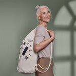 Eine lächelnde ältere Frau mit grauem Haar in einem Pferdeschwanz steht seitlich und hält eine weiße Liebscher & Bracht Faszien-Rollmassage-Set-Tasche mit einem minimalistischen Blattdesign und dem Text "Beweglichkeit & Entspannung" vor einem gedämpften grünen Hintergrund.