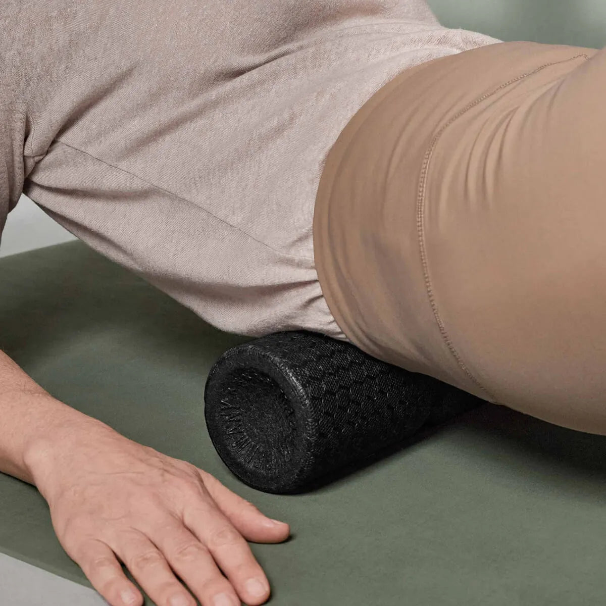 Eine Person benutzt eine schwarze Schaumstoffrolle von Liebscher & Bracht unter der Hüfte auf einer grünen Matte, trägt eine beige Hose und ein helles Hemd, inspiriert von "Rückenschmerzen verstehen und sich selbst davon befreien".