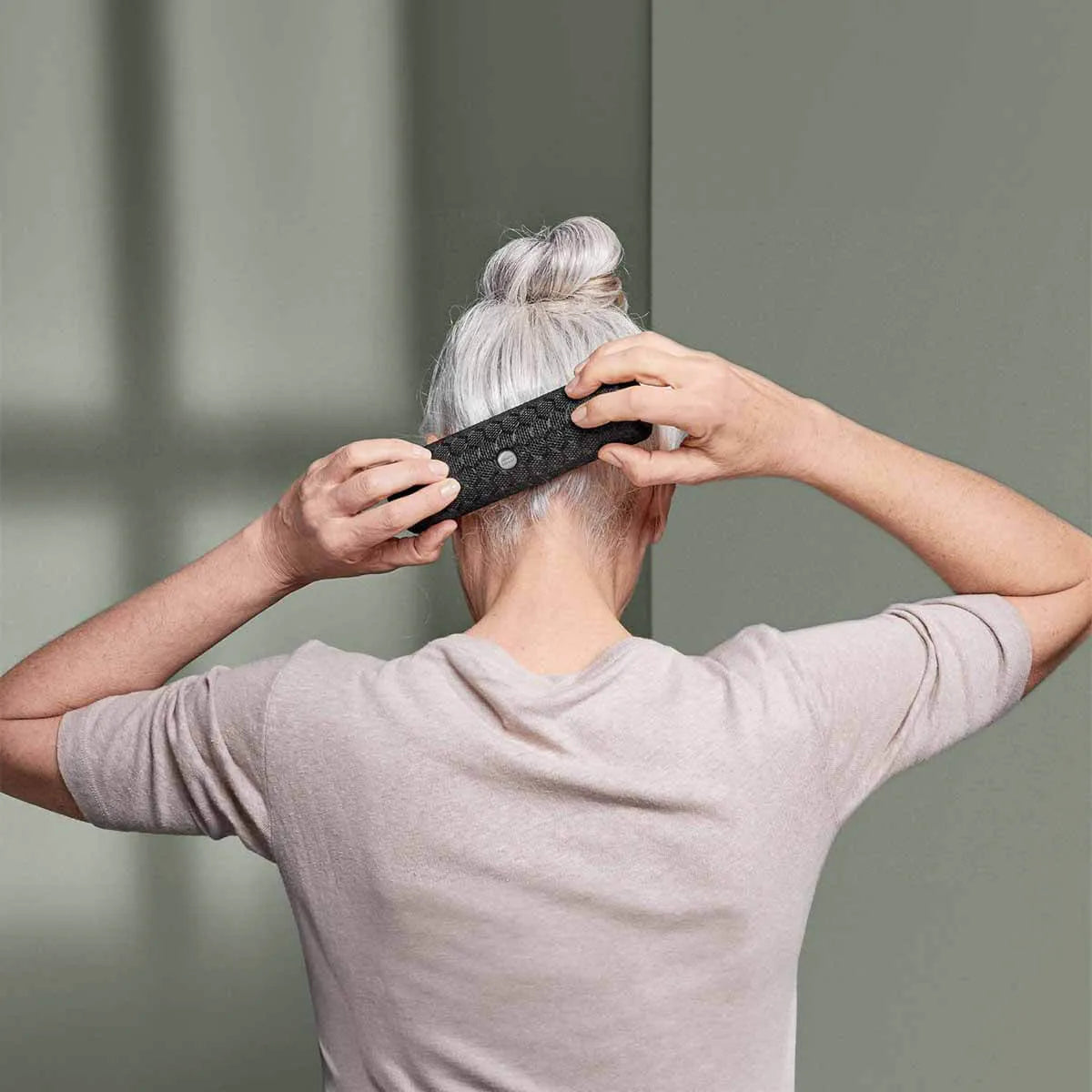 Eine Person mit grauem Haar in einem Dutt, die ein hellbeiges Hemd trägt, ist von hinten zu sehen, die das Liebscher & Bracht-Produkt "Rolle dich schmerzfrei" hinter ihrem Kopf vor einem gedämpften grünen Hintergrund hält.