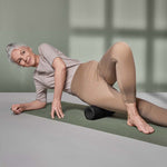 Eine grauhaarige Frau in beiger Sportkleidung nutzt das Faszien-Rollmassage-Set von Liebscher & Bracht auf ihrem äußeren Oberschenkel, während sie in einem sanft beleuchteten Raum auf einer Yogamatte liegt, um Entspannung und Beweglichkeit zu fördern.