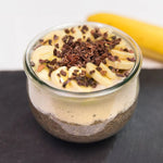 Ein Glasgefäß mit Chia-Pudding, Bananenscheiben und Kakaonibs - ein perfekter Intervallfasten-Snack von Liebscher & Bracht - präsentiert gesunde Ernährung. Im Hintergrund ruht eine verschwommene Banane auf einer dunklen Fläche.
