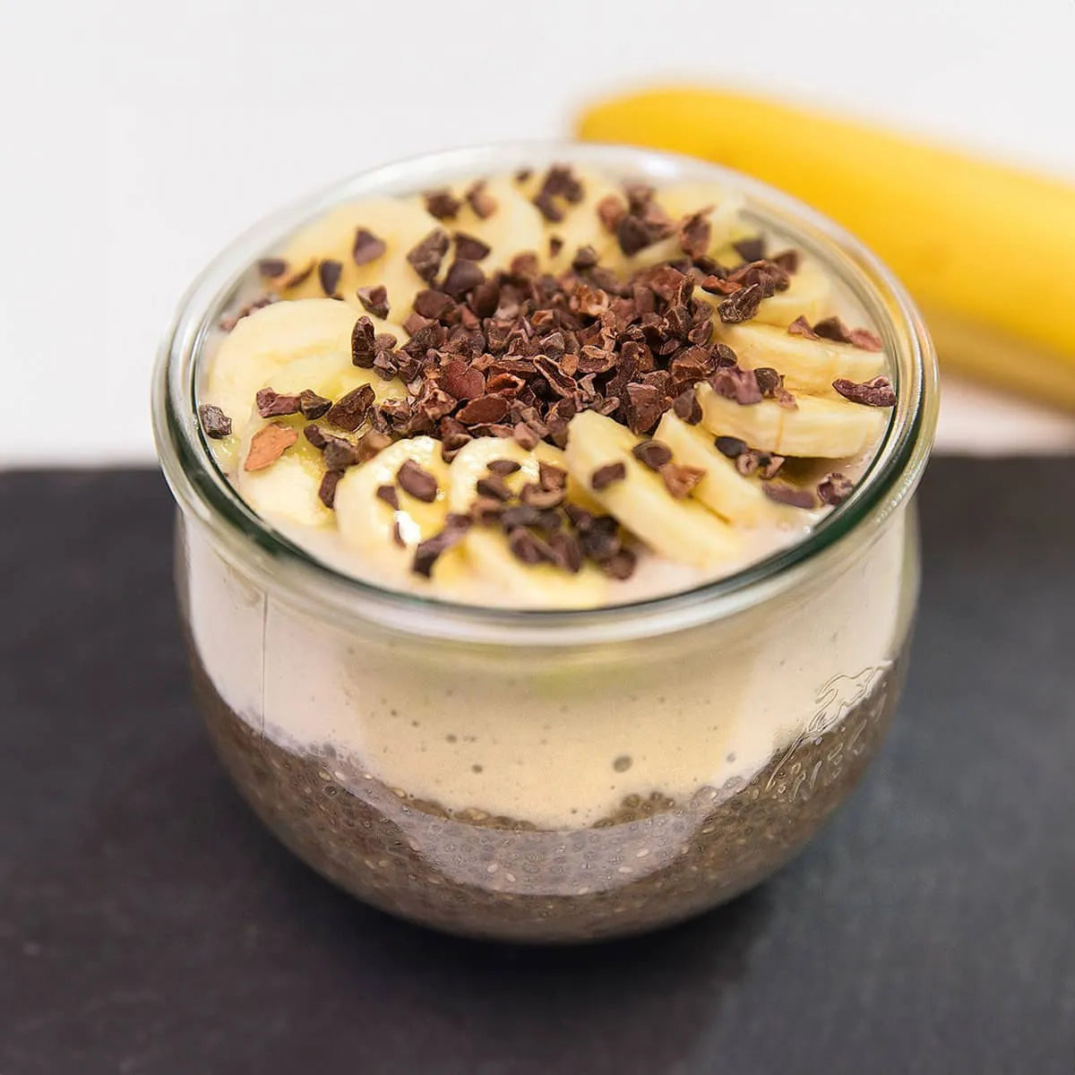 Ein Glasgefäß mit Chia-Pudding, Bananenscheiben und Kakaonibs - ein perfekter Intervallfasten-Snack von Liebscher & Bracht - präsentiert gesunde Ernährung. Im Hintergrund ruht eine verschwommene Banane auf einer dunklen Fläche.