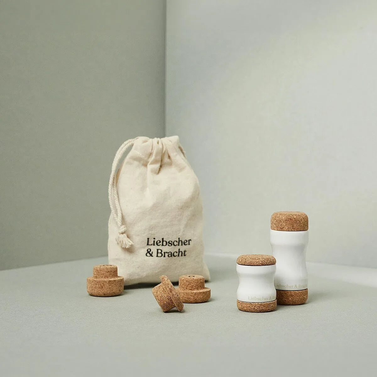 Ein Stoffbeutel mit der Aufschrift Liebscher & Bracht liegt neben dem Kieferretter, einem Set aus weißen Keramikwerkzeugen mit Korkplatten und Scheiben - ideal für Entspannung und Dehnübungen im Kieferbereich, angeordnet auf einer neutralen hellgrauen Fläche.