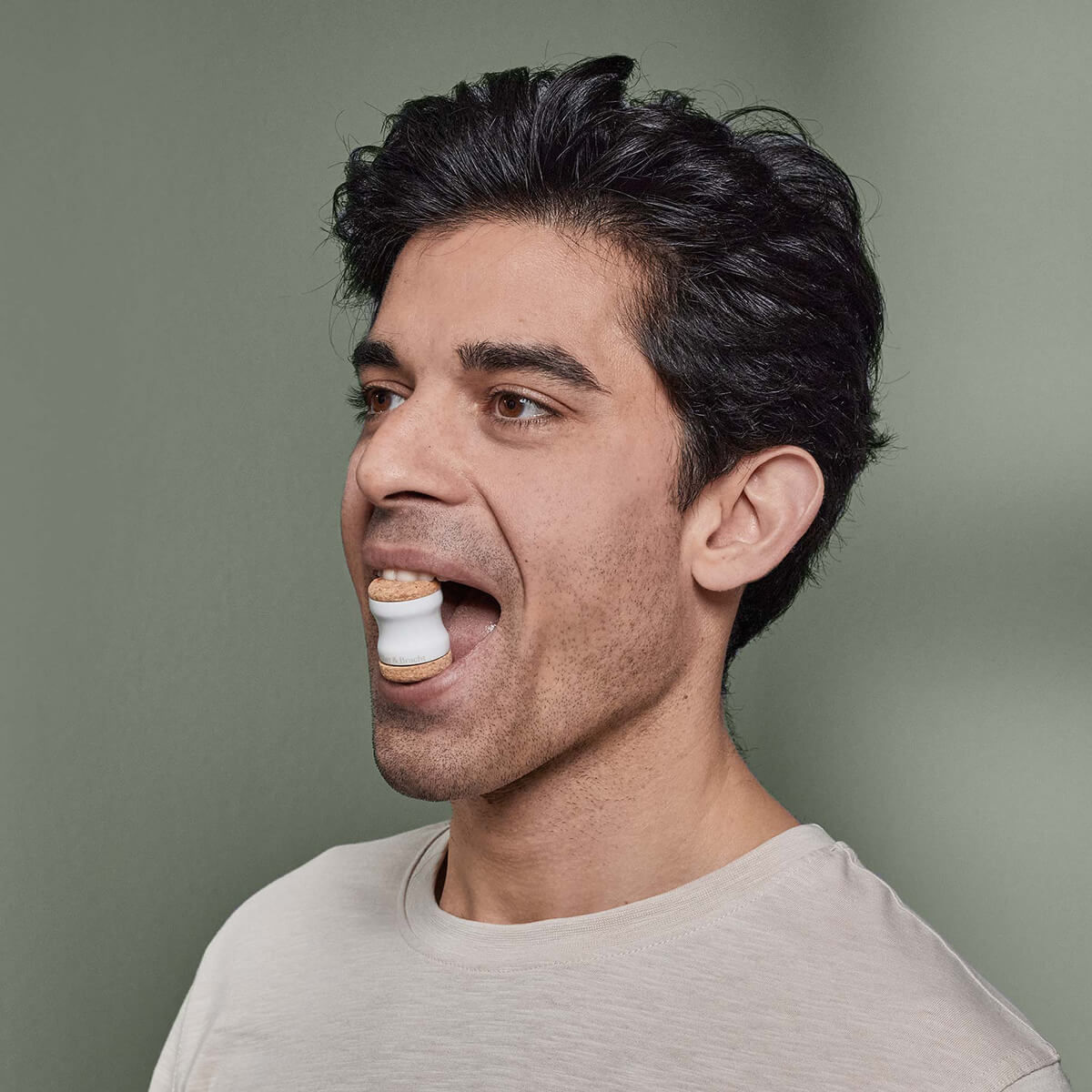 Ein dunkelhaariger Mann in einem hellen Hemd hält eine große weiße Der Kieferretter-Mütze von Liebscher & Bracht mit zwei Tabletten im offenen Mund, die auf Kieferentspannungs- und Dehnungsübungen vor einem schlichten grünen Hintergrund hinweisen.