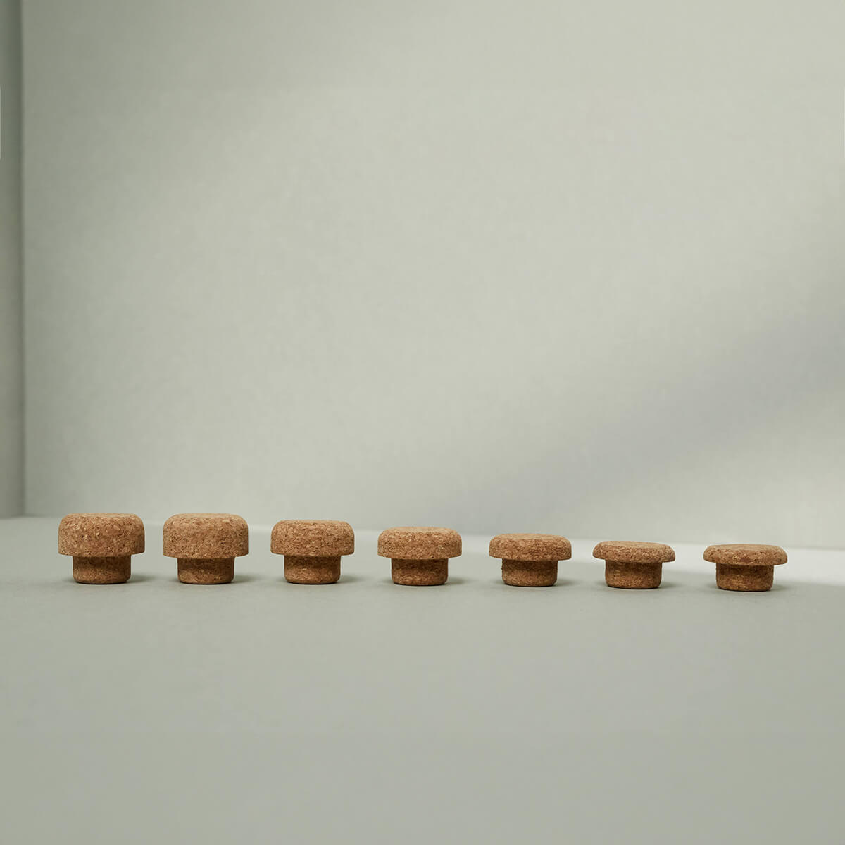 Sieben braune, unterschiedlich hohe Korken aus dem Korken-Set für den Kieferretter von Liebscher & Bracht sind auf einer glatten, hellgrauen Fläche vor einem schlichten hellgrauen Hintergrund aneinandergereiht.