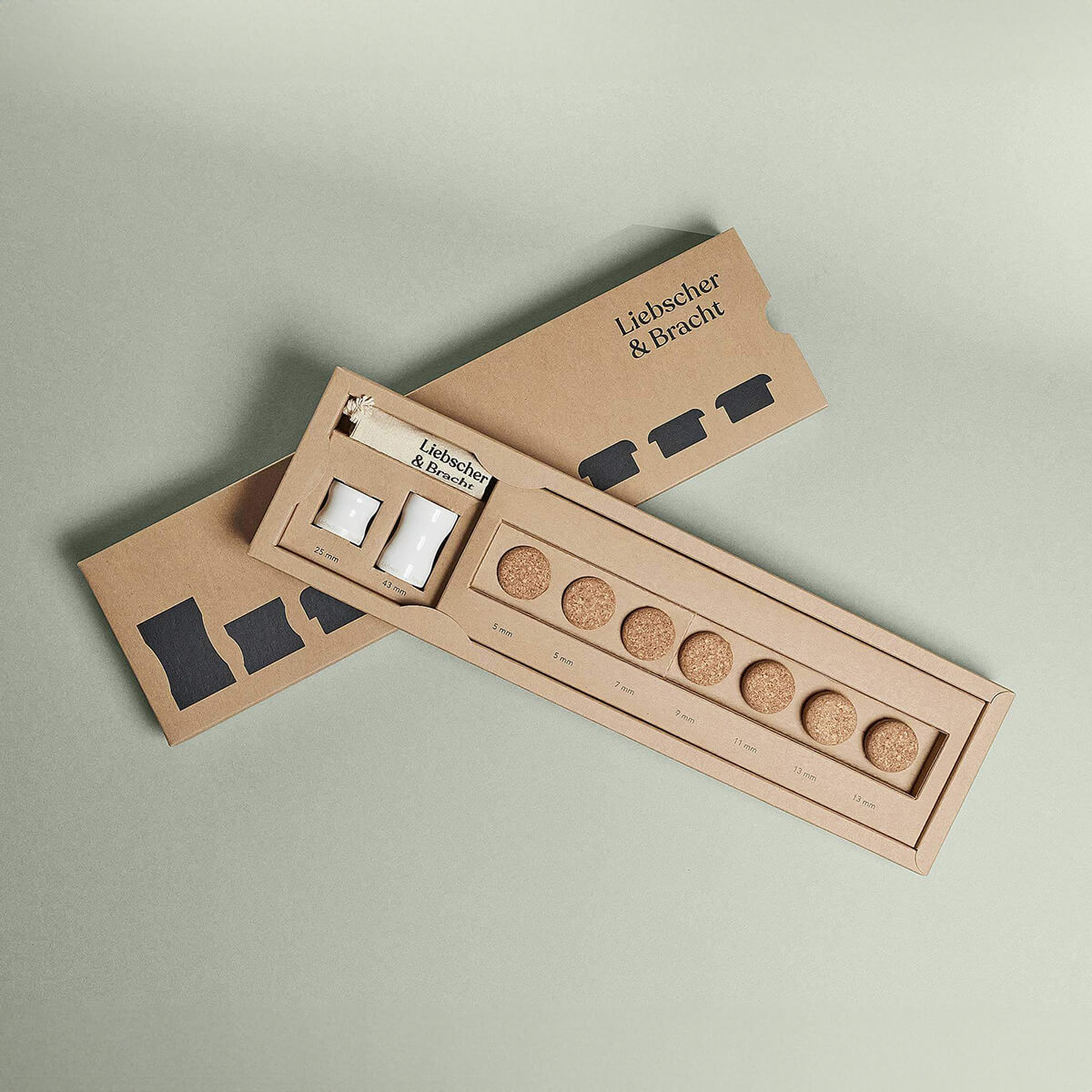 Das Produkt "Der Kieferretter" von Liebscher & Bracht wird in einer rechteckigen Schachtel mit Schiebedeckel geliefert, die acht runde Korkscheiben und zwei kleine weiße Schachteln enthält - perfekt für Entspannungs- und Kieferdehnungsübungen. Die Verpackung ist mit einem hellgrünen Etikett versehen.