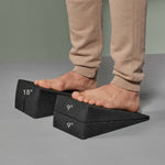 Eine Person in beiger Hose steht barfuß auf zwei gestapelten Schaumstoffkeilen "Der Knieretter mit Trittschlaufe" von Liebscher & Bracht, die jeweils mit einem Winkel von 18°, 9° und 9° gekennzeichnet sind und wahrscheinlich für Dehnungs- oder Fußstützübungen verwendet werden.