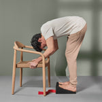 Eine Person steht auf einem schrägen Block, beugt sich mit leicht gebeugten Knien nach vorne und stützt sich mit den Armen auf einem Holzstuhl ab, während in der Nähe ein rotes Band auf dem Boden liegt.