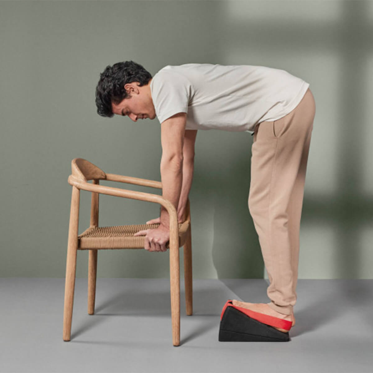 Eine Person in beiger Hose und weißem Hemd beugt sich nach vorne und stützt sich mit den Händen auf einen Holzstuhl, während sie auf einem Schaumstoffkeil steht und die Fersen anhebt.