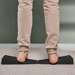 Eine Person in beiger Hose benutzt zwei schwarze Der Knieretter mit Trittschlaufe Schrägbretter von Liebscher & Bracht, um ihre Waden auf einem hellen Teppich zu dehnen, mit einer grünen Wand dahinter und der mitgelieferten Trittschlaufe in der Nähe.