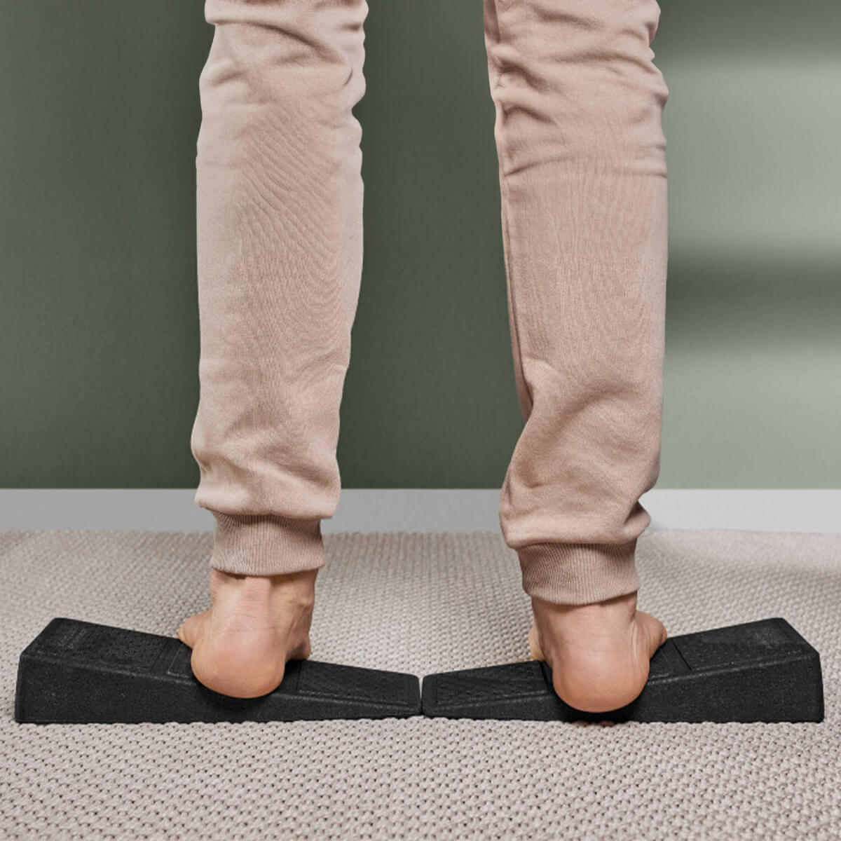 Eine Person in beiger Hose benutzt zwei schwarze Der Knieretter mit Trittschlaufe Schrägbretter von Liebscher & Bracht, um ihre Waden auf einem hellen Teppich zu dehnen, mit einer grünen Wand dahinter und der mitgelieferten Trittschlaufe in der Nähe.