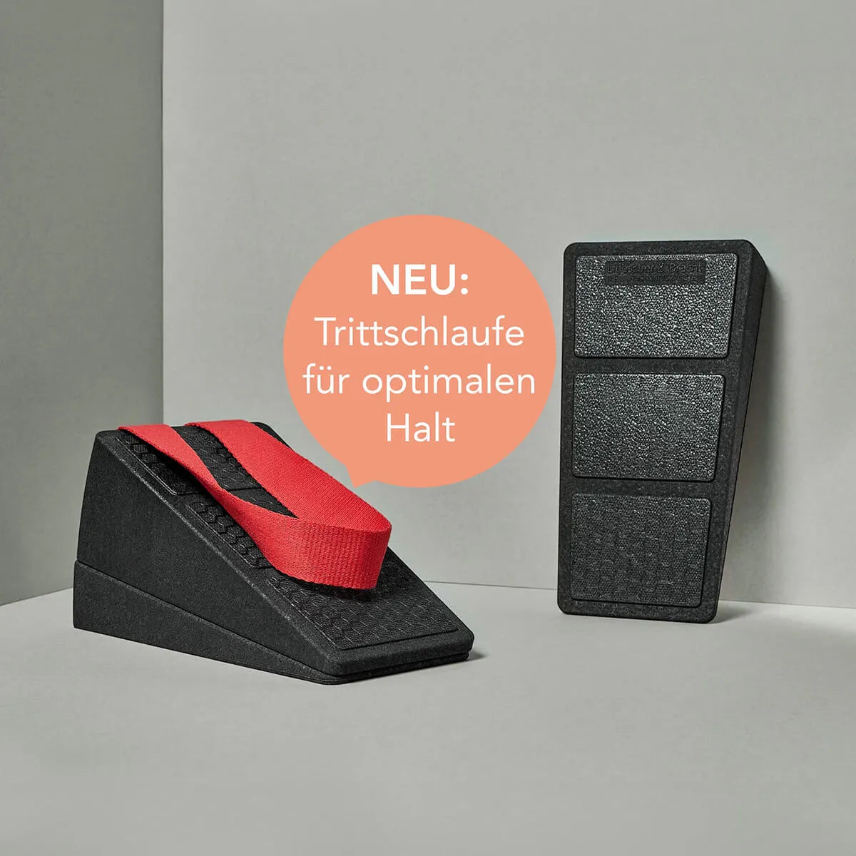 Zwei schwarze Schaumstoffkeile, "Der Knieretter mit Trittschlaufe" von Liebscher & Bracht, sind auf einer hellen Fläche abgebildet. In einem orangefarbenen Kreis steht "NEU: Trittschlaufe für optimalen Halt", was auf die neue Funktion für Dehnen und Knieretter-Übungen hinweist.