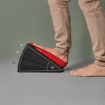 Jemand streckt seinen Fuß auf dem Liebscher & Bracht Dein Knie-Paket aus schwarzem Schaumstoff mit verstellbaren Winkeln (9°, 18°, 27°, 36°) und Trittschlaufe, in beiger Hose auf grauem Boden.