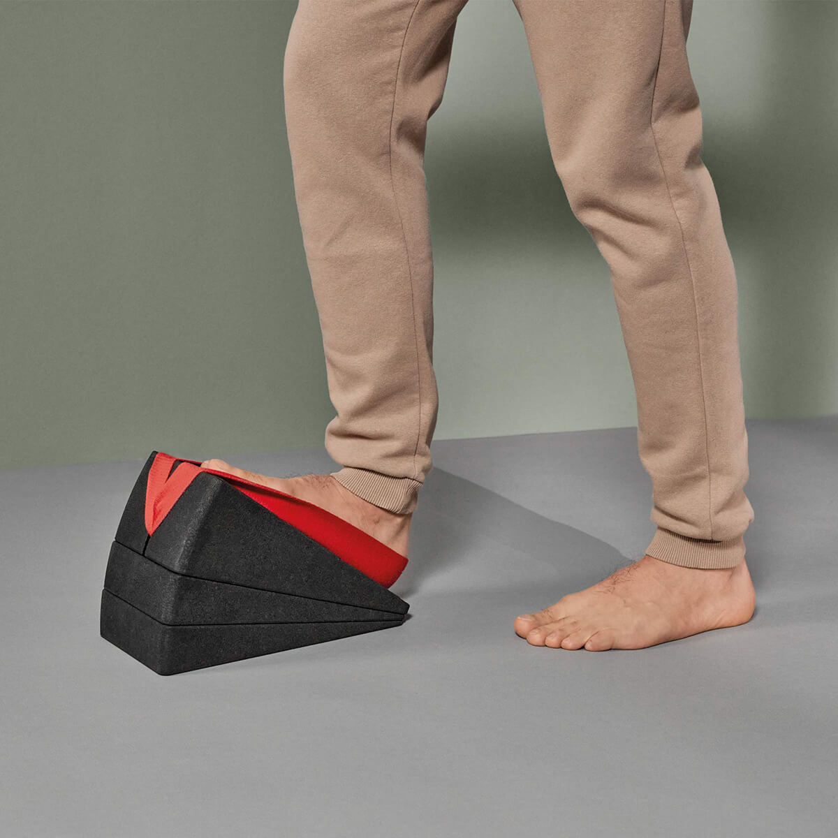 Eine Person in beiger Hose stellt einen Fuß auf die schwarz-rote Liebscher & Bracht Der Knieretter mit Trittschlaufe, während der andere nackte Fuß auf dem Boden steht und eine Dehnungs- oder Physiotherapieübung demonstriert.