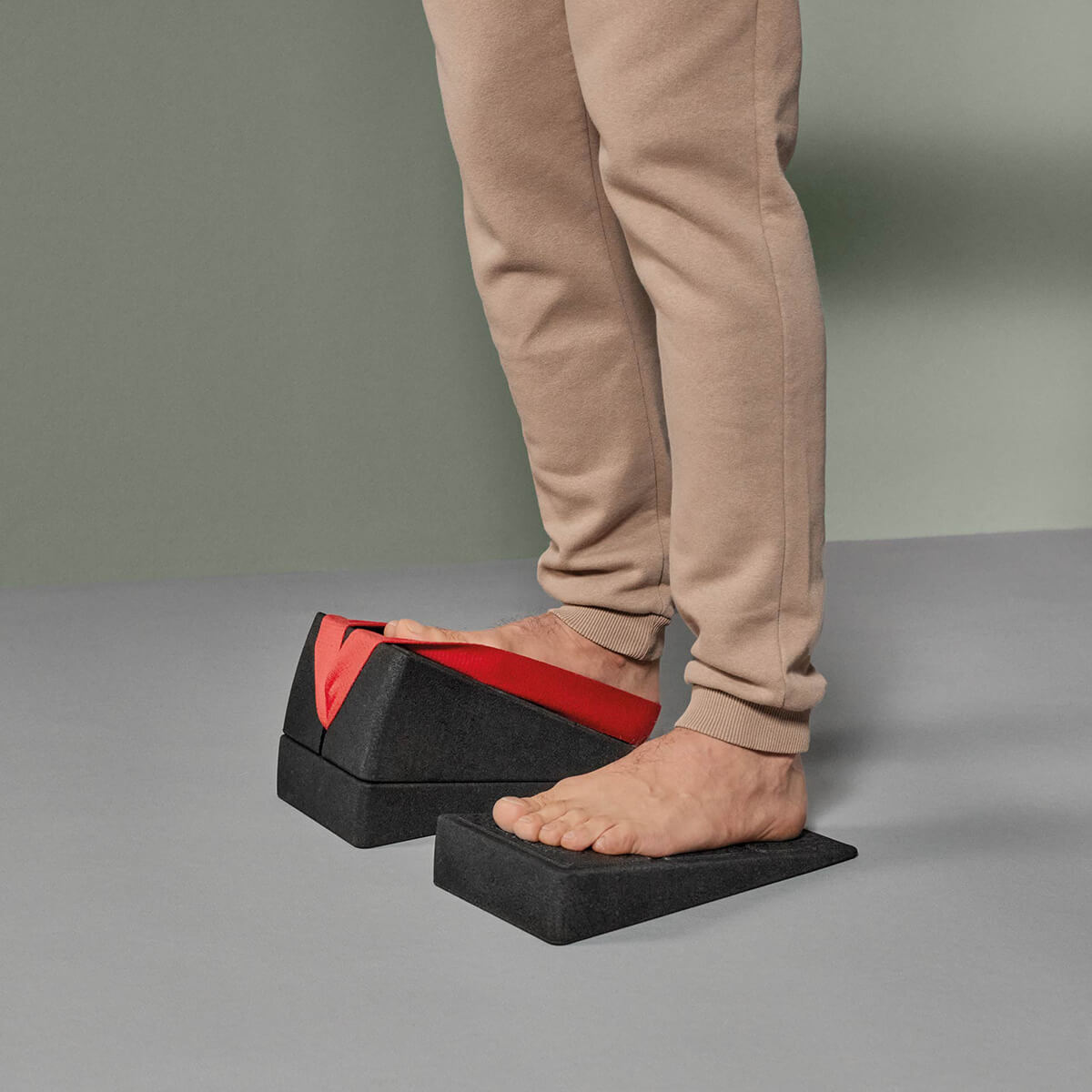 Eine Person in beiger Hose steht barfuß auf einem grauen Keil, während sie den Liebscher & Bracht Der Knieretter mit Trittschlaufe für Dehnübungen benutzt, vor einem grauen Hintergrund.