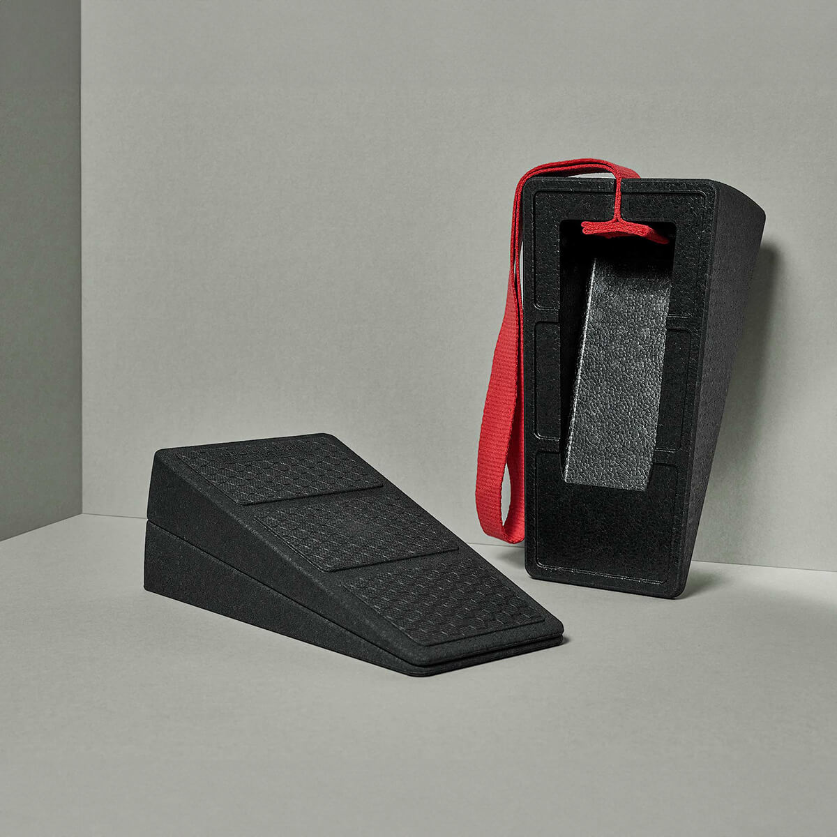 Abgebildet sind zwei schwarze Schaumstoffkeile "Liebscher & Bracht Der Knieretter mit Trittschlaufe", ein flacher und ein aufrechter, an denen ein roter Fußriemen befestigt ist. Der Hintergrund besteht aus einer neutralen, hellgrauen Fläche und Wand.