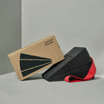 Eine Schachtel mit der Aufschrift Liebscher & Bracht steht aufrecht neben zwei gestapelten schwarzen Schaumstoffkeilen mit einem roten Trittschlaufe-Band, auf dem Der Knieretter mit Trittschlaufe abgebildet ist, alles vor einem neutralgrauen Hintergrund.