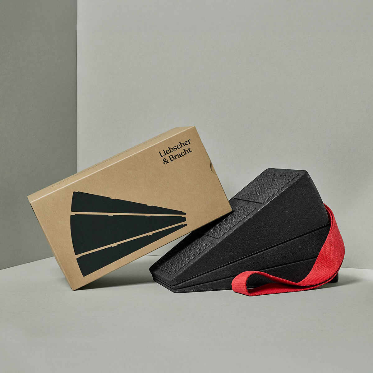 Eine Schachtel mit der Aufschrift Liebscher & Bracht steht aufrecht neben zwei gestapelten schwarzen Schaumstoffkeilen mit einem roten Trittschlaufe-Band, auf dem Der Knieretter mit Trittschlaufe abgebildet ist, alles vor einem neutralgrauen Hintergrund.