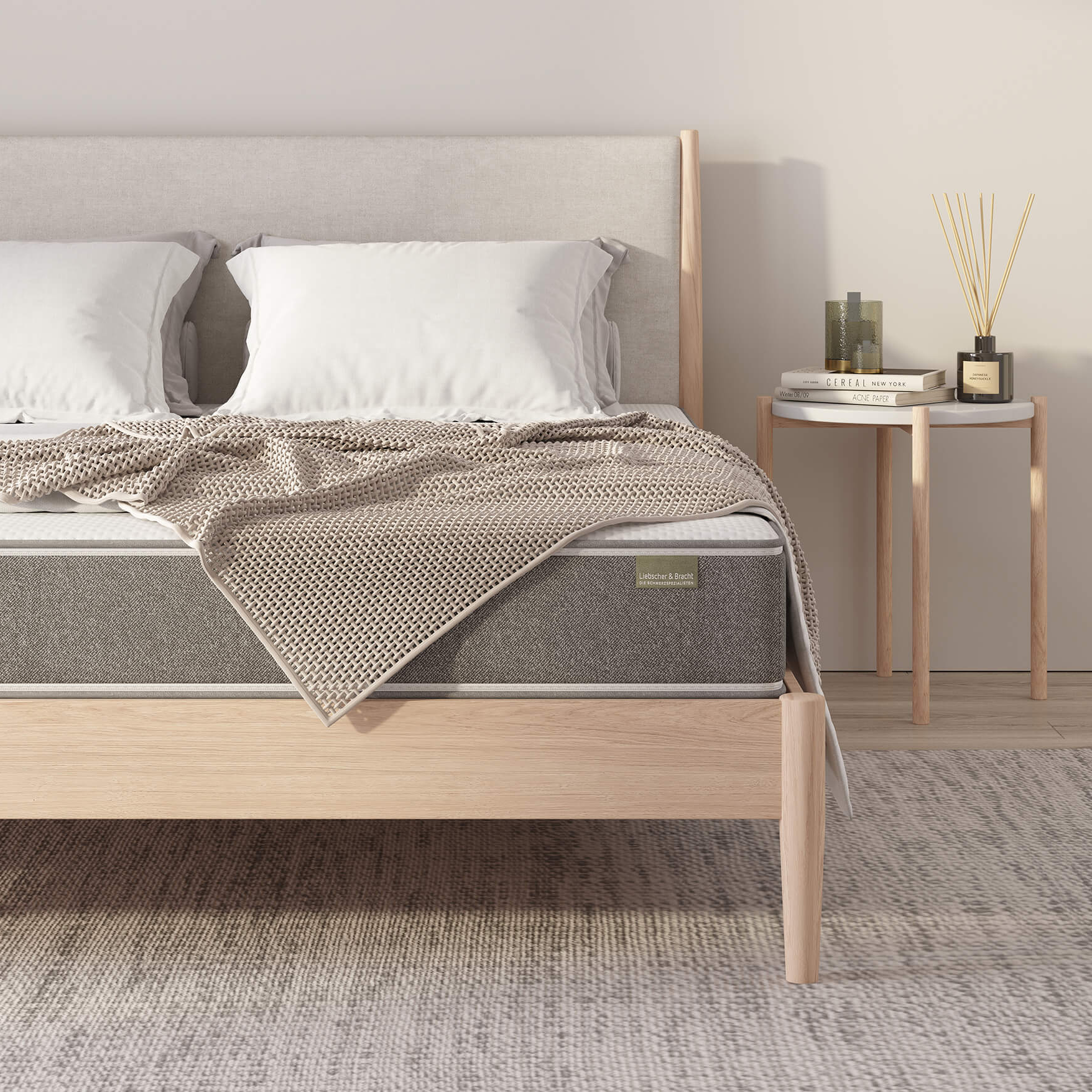Ein ordentlich gemachtes Bett mit weißen Kissen, einer beigen Decke und einer grauen Matratze steht auf einem hellen Holzrahmen. Daneben steht ein kleiner runder Beistelltisch mit Büchern, einem Diffusor und einer Kerze darauf.