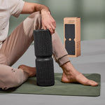 Eine Person in beiger Hose sitzt auf einer Yogamatte und hält die Liebscher & Bracht MAXI-Faszienrolle mit Wirbelsäulen-Entlastung-ideal für große Körperflächen. Im Hintergrund ist ein brauner Markenkarton zu sehen.