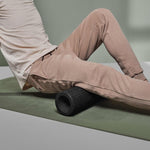 Eine Person in beiger Hose und weißem Hemd benutzt die MAXI-Faszienrolle mit Wirbelsäulen-Entlastung von Liebscher & Bracht, ideal für große Muskelpartien, auf dem Oberschenkel, während sie auf einer grünen Übungsmatte sitzt.