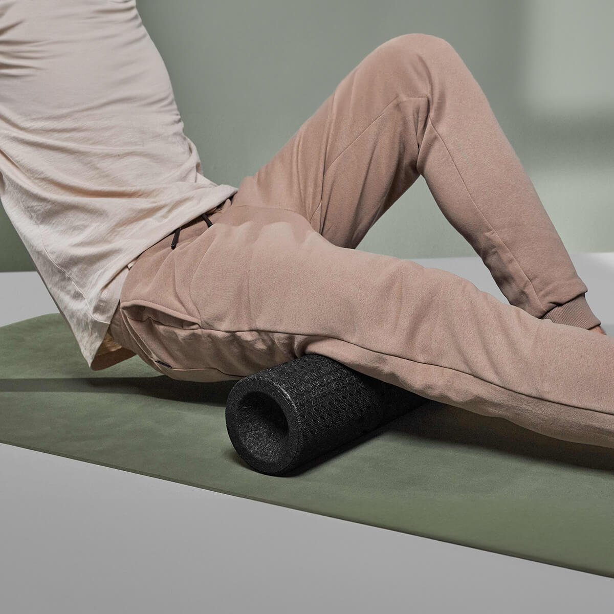 Eine Person in beiger Hose und weißem Hemd benutzt die MAXI-Faszienrolle mit Wirbelsäulen-Entlastung von Liebscher & Bracht, ideal für große Muskelpartien, auf dem Oberschenkel, während sie auf einer grünen Übungsmatte sitzt.