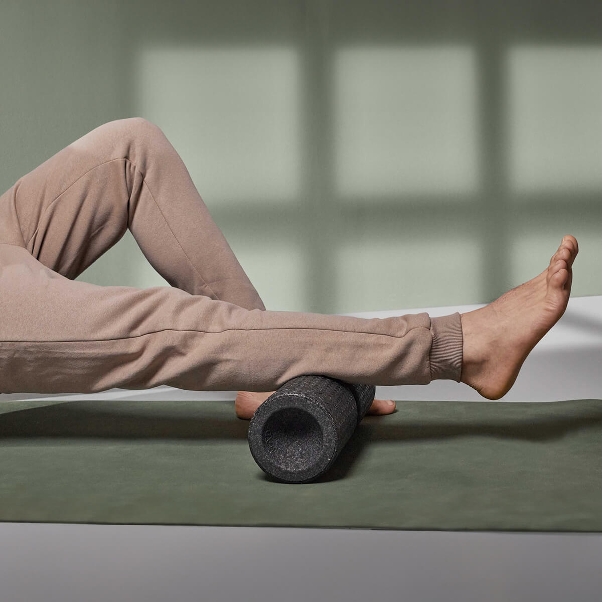 Eine Person in hellbrauner Jogginghose benutzt die Liebscher & Bracht MAXI-Faszienrolle mit Wirbelsäulen-Entlastung unter der Wade auf einer grünen Matte in einem sonnenbeschienenen Raum, ideal für große Körperflächen oder zur gezielten Behandlung der Wirbelsäule.
