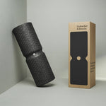 Die Liebscher & Bracht MAXI-Faszienrolle mit Wirbelsäulen-Entlastung, eine strukturierte schwarze Schaumstoffrolle für große Körperflächen, lehnt an einer hellgrauen Wand neben ihrer braunen Schachtel mit dem Markenlogo und einem Ausschnitt der Rollenform.