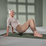 Eine ältere Frau in Trainingskleidung lächelt, während sie auf einer Yogamatte sitzt und das Liebscher & Bracht Unsere Empfehlung | Faszien-Set + Übungs-Schlaufe unter ihrem unteren Rücken für Dehnungsübungen und Selbstmassage verwendet.