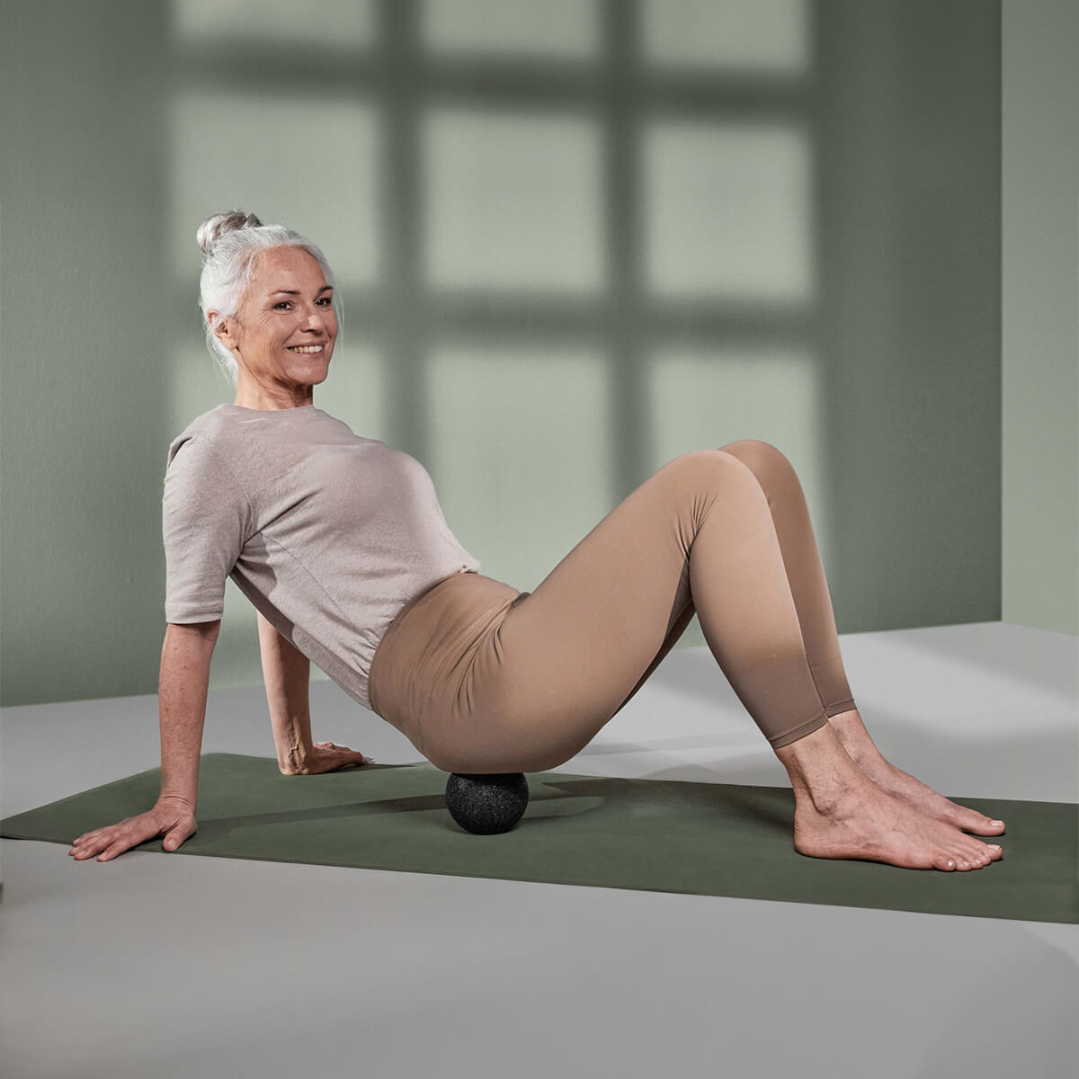 Eine ältere Frau in Trainingskleidung lächelt, während sie auf einer Yogamatte sitzt und das Liebscher & Bracht Unsere Empfehlung | Faszien-Set + Übungs-Schlaufe unter ihrem unteren Rücken für Dehnungsübungen und Selbstmassage verwendet.