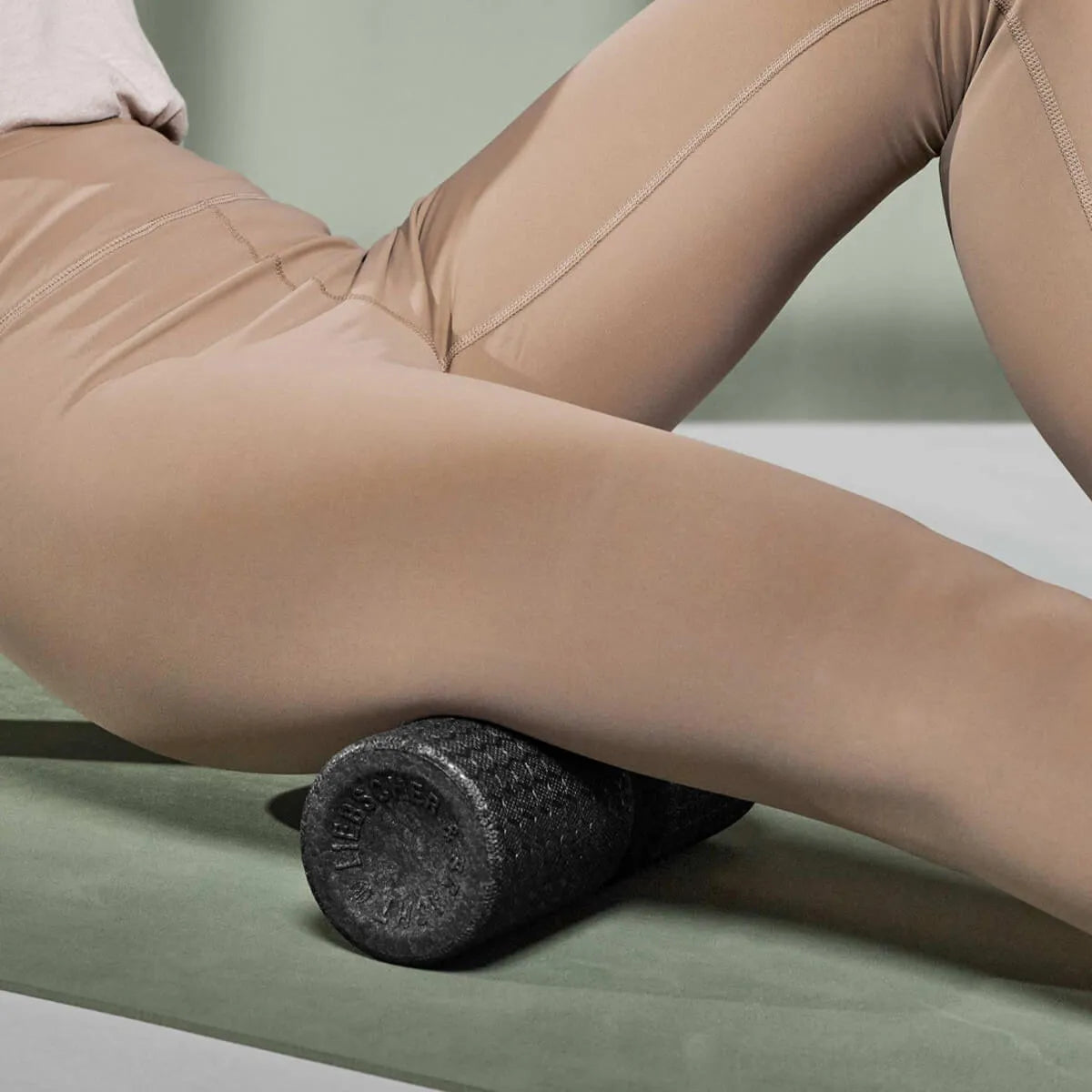 Eine Person in beiger Leggings benutzt die schwarze Schaumstoffrolle "Rolle dich schmerzfrei" von Liebscher & Bracht unter dem Oberschenkel auf einer grünen Matte und übt sich in der Faszienentspannung.