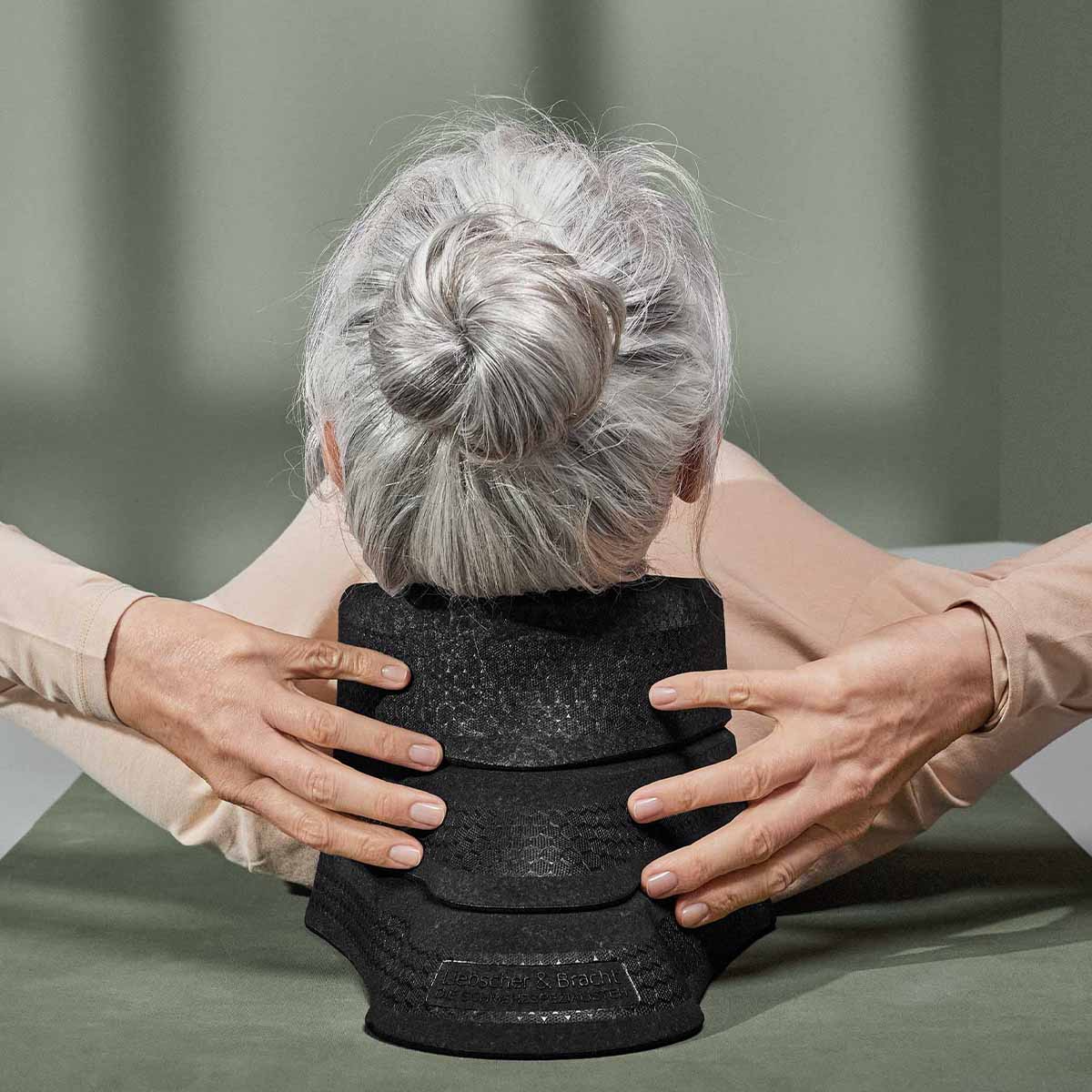 Eine ältere Frau mit grauem Haar in einem Dutt liegt auf dem Rücken und benutzt Der Nackenretter von Liebscher & Bracht für Entspannungsübungen. Sie ruht sich auf einer grün-grauen Fläche aus und stützt das Gerät mit den Händen.