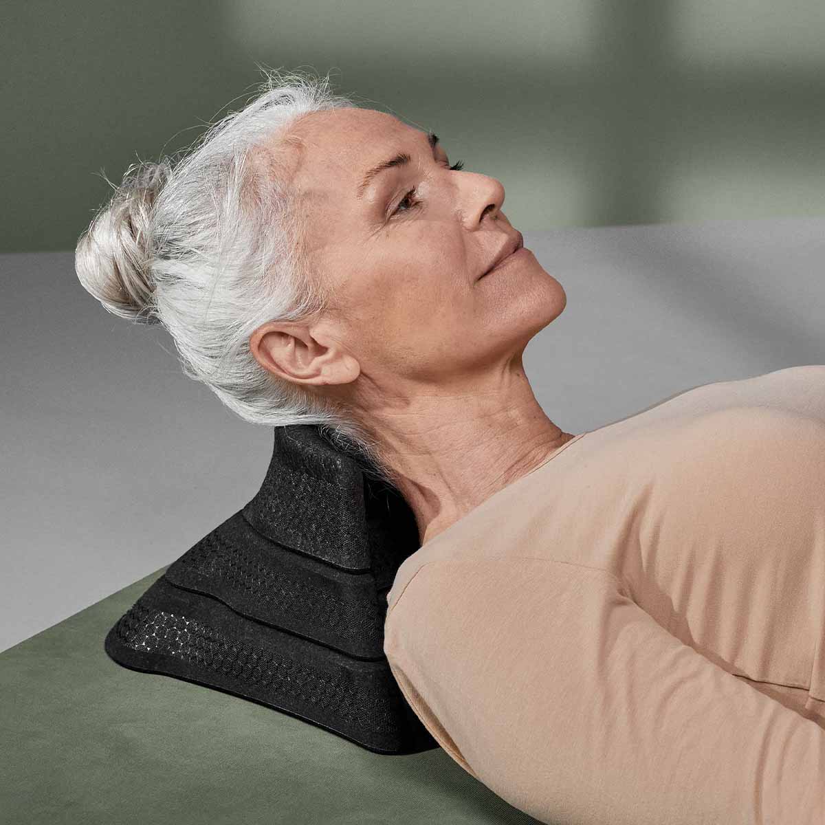 Eine ältere Frau mit grauem Haar in einem Dutt liegt auf dem Rücken und scheint sich bei Entspannungsübungen zu entspannen, während sie ihren Nacken auf Der Nackenretter von Liebscher & Bracht auf einer grün-grauen Fläche abstützt.