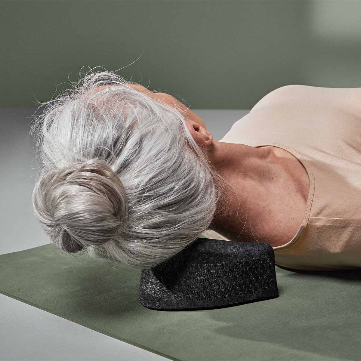 Eine Person mit grauem Haar in einem Dutt liegt auf dem Rücken auf einer Yogamatte und benutzt Der Nackenretter von Liebscher & Bracht, ein strukturiertes schwarzes Nackenstützkissen für Dehnungs- und Entspannungsübungen.